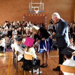 Starsza kobieta i mężczyzna w średnim wieku przekazujący dzieciom upominki na sali gimnastycznej. W tle siedzący na krzesłach i ławkach dorośli i dzieci.