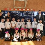 Grupa dzieci odświętnie ubranych pozujących do zdjęcia na sali gimnastycznej. Stoi z nimi mężczyzna w garniturze oraz dwie Panie.