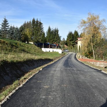 Szeroka droga ze świeżo położonym asfaltem. Nieopodal krzewy i trawa oraz kiszonki siana. Panuje słoneczna pogoda.