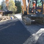 Koparka pracująca na drodze asfaltowej. Trwają pacę nad wykonaniem wjazdu na działkę prywatną. Panuje słoneczna pogoda. W tle widać krzewy i drzewa.