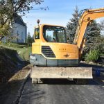 Szeroka droga ze świeżo położonym asfaltem. Na pierwszym planie koparka. W tle widać mały, biały kościołek.