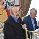 Dwóch starszych mężczyzn siedzących przy okrągłym stole. Jeden z nich przemawia za pomocą mikrofonu. Z tyłu widać duży baner i zasłonę.