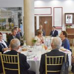 Duża grupa elegancko ubranych osób różnej płci zasiada przy okrągłych stołach na rozległej sali bankietowej.