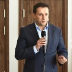 Mlody mężczyzna w garniturze przemawia za pomocą mikrofonu. Z tyłu widać drzwi oraz ścianę.