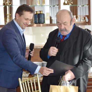 Dwóch mężczyzn w garniturach. Młodszy przekazuje starszemu upominki. Z tyłu widzimy półkę oraz blat.