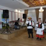 Grupa osob starszych w strojach ludowych wykonujący utwór muzyczny. Znajdują się na dużej sali. Z ich lewej strony znajduje się podwyższenie z banerami oraz sprzętem muzycznym.