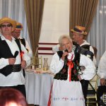 Grupa osob starszych w strojach ludowych wykonujący utwór muzyczny. Z tyłu widać stoły oraz ozdobne zasłony.