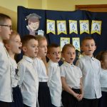 Grupa dzieci ubrane w odświętnie stroje biało czarne. Za nimi tablica granatowa.