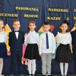 Grupa dzieci ubrane w odświętnie stroje biało czarne. Za nimi tablica granatowa.