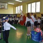 Grupa dzieci ubrana w odświętne stroje, składa pasowanie, przed nim stoją ludzie z sztanadrem.