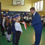 Grupa dzieci ubrana w odświętne stroje, Mężczyzna w garniturze pasuje dzieci ołowkiem.