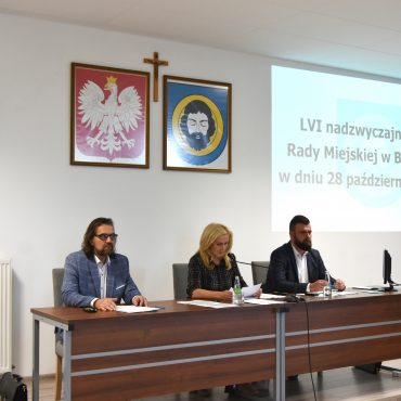 Dwóch mężczyzn i kobieta w średnim wieku siedzący przy długim stole. Wszyscy są odświętnie ubrani. Na ścianie widać herby, krzyż i obraz z wyświetlacza.