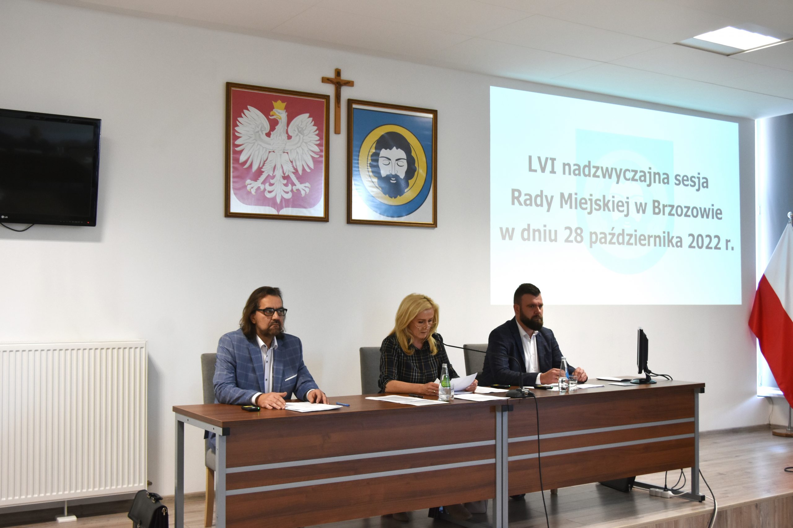 Dwóch mężczyzn i kobieta w średnim wieku siedzący przy długim stole. Wszyscy są odświętnie ubrani. Na ścianie widać herby, krzyż i obraz z wyświetlacza.