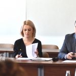 Mężczyzna i kobieta siedzący przy długim biurku na rozległej dobrze oświetlonej sali. Mężczyzna spogląda do przodu kobieta zaś uważnie przegląda dokument.