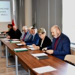 Osoby w średnim i starszym wieku obojga płci siedzący przy stolach na dużej sali. Na stołach leży mikrofon i kartki. Na ścianie widać obraz z wyświetlacza.