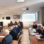 Osoby w średnim i starszym wieku obojga płci siedzący przy stolach na dużej sali. Na stołach leży mikrofon i kartki. Na ścianie widać obraz z wyświetlacza, herby i krzyż.