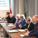 Osoby w średnim i starszym wieku obojga płci siedzący przy stolach na dużej sali. Na stołach leży mikrofon i kartki. Na ścianie widać obraz z wyświetlacza.