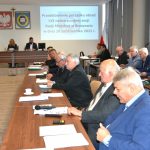 Osoby w średnim i starszym wieku obojga płci siedzący przy stolach na dużej sali. Na stołach leży mikrofon i kartki. Na ścianie widać obraz z wyświetlacza, herby i krzyż.