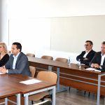 Trzech mężczyzn i kobieta zasiadający przy stolach. Wszyscy są odświętnie ubrani. Na stolach lezą kartki papieru i mikrofon.