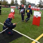 Grupa strażaków ćwiczących na stadionie. Jeden celuje wężem strażackim do tarczy.