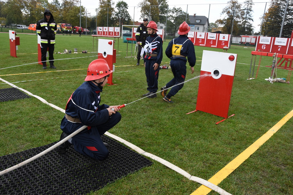 Grupa strażaków ćwiczących na stadionie. Jeden celuje wężem strażackim do tarczy.