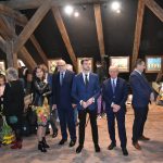 Grupa elegancko ubranych osób (głównie mężczyzn)pozuje do zdjęcia. W tle ściana poddasza.