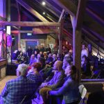 Grupa osób w różnym wieku obserwująca występ muzyczny na wielkim poddaszu.