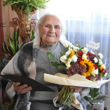 Starsza kobieta z siwymi włosami siedzi na krześle okryta szalikiem, trzyma w rękach bukiet kwiatów i teczkę z życzeniami.