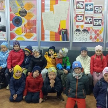 Grupa dzieci ubrana w kurtki i czapki siedzi na krzesełkach w sali teatralnej. Za nimi plakaty.