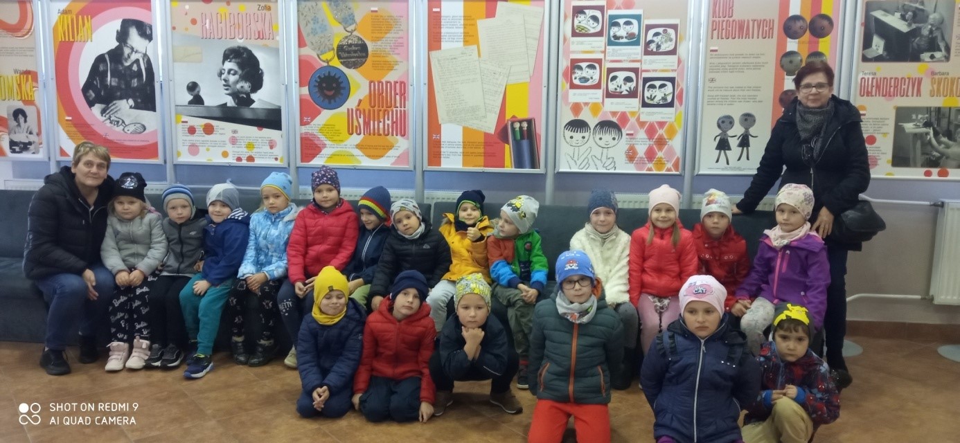 Grupa dzieci ubrana w kurtki i czapki siedzi na krzesełkach w sali teatralnej. Za nimi plakaty.