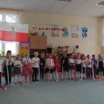 Grupa dzieci ubrana w odświętne stroje stoi na dywanie w sali szkolnej i śpiewa piosenkę. W rękach dzieci trzymają flagi biało czerwone. Za nimi na ścianie wisi duża flaga biało czerwona.