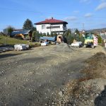 Remontowany plac na którym widać sprzęt i maszyny budowlane. W tle można dostrzec ulicę, domy oraz drzewa i krzewy. Panuje piękna słoneczna pogoda.