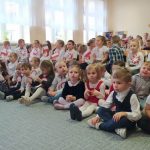 Grupa dzieci ubrana w odświętne stroje, siedzią na dywanie w sali szkolnej i oglądają przedstawienie. Niektóre dzieci w rękach trzymają małe flagi państwowe.