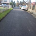 Droga z nowo położonym asfaltem. W tle widac budynki mieszkalne i samochody.