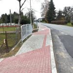 Chodnik z kostek brukowych oraz droga asfaltowa. W tle lampy, domy, i krzewy.