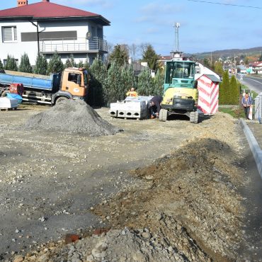 Remontowany plac na którym widać sprzęt i maszyny budowlane. W tle można dostrzec ulicę, domy oraz drzewa i krzewy. Panuje piękna słoneczna pogoda.