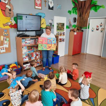 Grupa dzieci siedzi na dywanie w sali przedszkolnej. Przed nimi stoi młoda kobieta o krótkich włosach i trzyma w rękach plakat.
