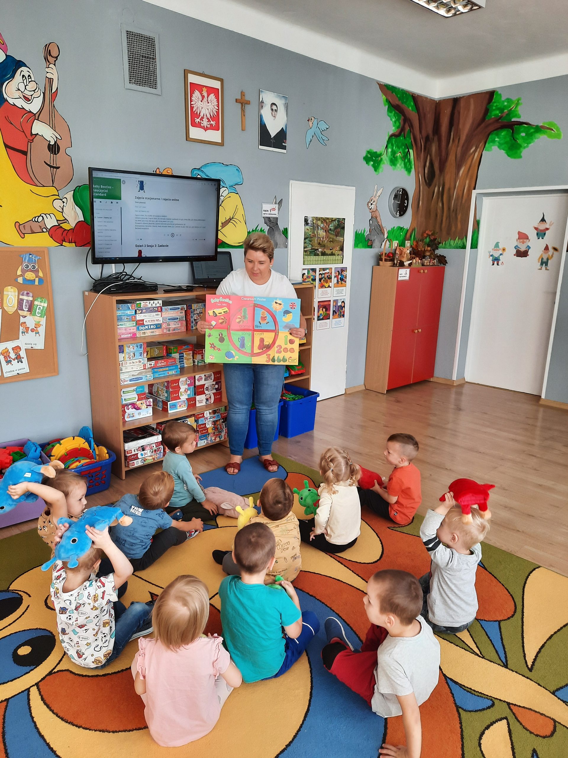 Grupa dzieci siedzi na dywanie w sali przedszkolnej. Przed nimi stoi młoda kobieta o krótkich włosach i trzyma w rękach plakat.