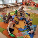 Grupa dzieci siedzi na dywanie kolorowym w przedszkolu, dzieci trzymają w rękach maskotki, za nimi ustawione są małe krzesełka i stoliki.
