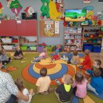 Grupa dzieci siedzi na dywanie w sali przedszkolnej i ogląda bajki na telewizorze.