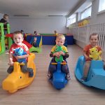 Tzrech chłopców siedzi na plastikowych konikach i trzymają w rękach maskotki. Za nimi znajduje się basen z kulkami.