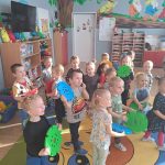 Grupa dzieci tańczy w kolowerj sali pzredszkolnej, trzymając w rękach plasitkowe kółka.