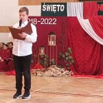 Młody chłopak ubrany w białą koszulę i czarne spodnie, trzyma w rękach czerwoną książkę i śpiewa do mikrofonu. Za nim wystojona czerowna tablica.