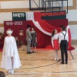 Na pierwszym planie dziewczynka ubrana na biało śpiewa pieśń, a za nią dwie pary tańczą. W dalszym planie wystrojona tablica czerwona.