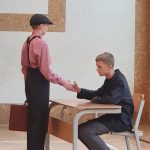 Młody chłopak ubrany w czerwoną koszulę i czapkę na głowie wita się z młodym chłopakiem ubranym w czarną bluzę, siedzi przy stoliku.