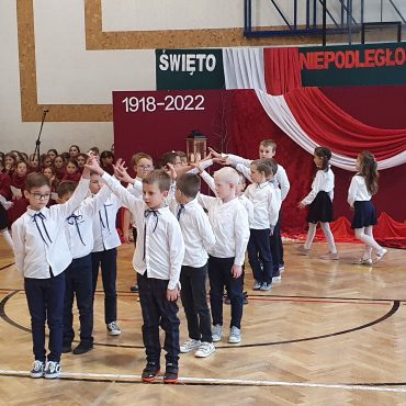 Grupa dzieci ubranych w odświętne stroje, białe koszule i ciemne spódnice, spodnie tancza poloneza na sali gimnastycznej.