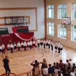 Grupa dzieci ubrana w odświętne stroje stoi na sali gimnastycznej i bierze udział w uroczystej akademii.