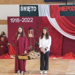 Dwie dziewczyny jedna ubrana w długą czerwoną szatę, a druga w białą koszule i czarne spodnie recytują wiersze na sali gimnastycznej.