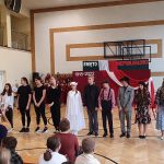 Grupa młodzieży przebrani w różne stroje [przedstawiają na sali gimnastyczną spektakl przed widownią.