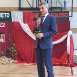 Mężczyzna w średnim wieku trzymający czerwoną różę w ręku ubrany w granatowy garnitur przemawia do mikrofonu na sali gimnastycznej.